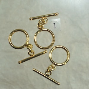 May include: Three gold-colored toggle clasps. Each clasp has a circular ring and a straight bar with rounded ends. The clasps are designed for jewelry making and are displayed on a light background.