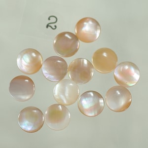 May include: A collection of round, iridescent, mother-of-pearl shell beads. The beads are a soft peach color with a subtle sheen, and are arranged on a light background. The number "2" is visible in the upper left corner.