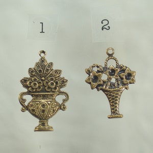Può includere: Due ciondoli antichi in metallo color oro. Un ciondolo è un vaso decorativo con fiori. L'altro ciondolo è un cestino di fiori. Entrambi i ciondoli hanno un anello nella parte superiore per attaccarli a una collana o un braccialetto.
