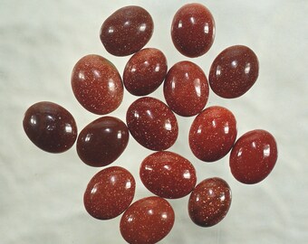 May include: A collection of oval-shaped, polished gemstones in shades of reddish-brown. The stones have a speckled, glittery appearance, with some appearing darker than others. The gemstones are arranged on a light-colored surface.