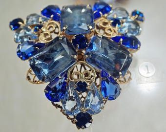 Peut inclure: Broche vintage ornée de pierres de verre bleues et bleu clair taillées en facettes de différentes formes. Les pierres sont serties dans un cadre en métal filigrané doré, créant un accessoire étincelant et accrocheur.