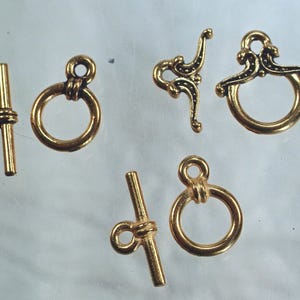 May include: An assortment of gold-colored jewelry clasps and toggle clasps. The clasps include a variety of ring and bar designs, with some featuring decorative embellishments. The pieces are arranged against a light background.