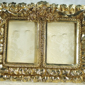 May include: A gold-coloured double picture frame with an ornate design. The frame has a ruffled edge, rope-like trim, and floral accents. The interior displays a cream-coloured fabric.