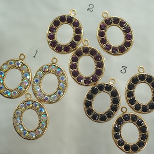 Op de afbeelding: Ovale hangers van goudkleurig metaal, elk versierd met een ring van sprankelende edelstenen. De edelstenen zijn in drie kleuren: iriserend, dieppaars en zwart. Elke hanger heeft een kleine lus aan de bovenkant om aan een ketting of andere sieraden te bevestigen.