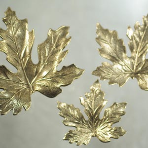 May include: Three gold-coloured maple leaf decorations. The leaves have detailed veining and a textured surface. The leaves are in different sizes and arranged on a reflective surface.