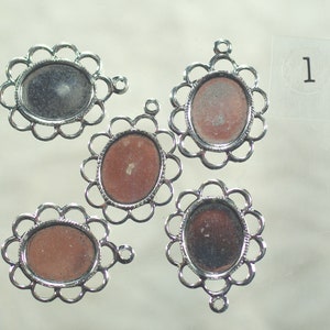 May include: Five silver-colored oval pendant trays with scalloped edges. The trays have a loop at the top for hanging and a recessed area to hold a cabochon. One tray has the number "1" on a sticker.