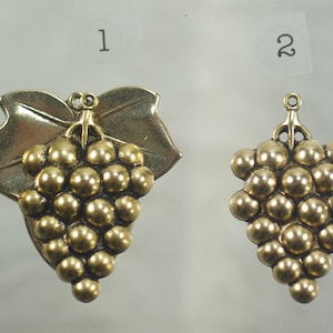 May include: Two gold-toned grape cluster charms. One charm features a leaf and a cluster of round grapes. The other charm is a smaller cluster of grapes. Both charms have a loop at the top for hanging.