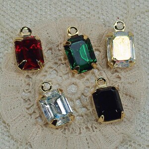 Puede incluir: Cinco colgantes rectangulares de piedras preciosas en rojo, verde, transparente, iridiscente y negro. Cada gema está engastada en un marco de metal dorado con un pequeño lazo en la parte superior para sujetar. Los colgantes se muestran sobre una superficie de encaje color crema.
