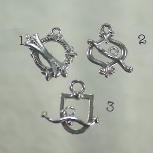 Könnte beinhalten: Drei silberfarbene Knebelverschlüsse für die Schmuckherstellung. Jeder Verschluss hat ein anderes Design: ein dekorativer Kreis, eine stilisierte Acht und eine Schildform. Die Verschlüsse werden vor einem hellen Hintergrund gezeigt.