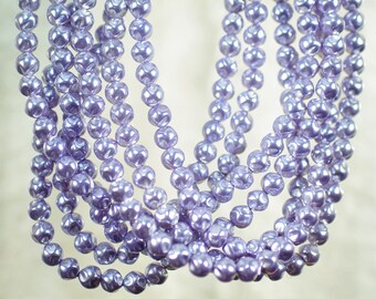 May include: Close-up of multiple strands of lavender-colored beaded necklaces. The beads have a textured, patterned surface and a glossy finish. The necklaces are arranged in a cascading, layered design, creating a visually appealing display of jewelry.