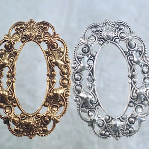 May include: Two ornate, oval-shaped frames. One is gold-toned, the other silver-toned. Both feature intricate floral and scrollwork designs with small bird-like figures. The frames are likely for jewelry or decorative purposes.