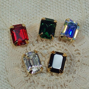 Puede incluir: Cinco cabujones de piedras preciosas rectangulares en rojo, verde, transparente, iridiscente y negro, cada uno engastado en un marco de metal dorado. Las piedras preciosas se muestran sobre una superficie texturizada de color crema, mostrando sus colores vibrantes.