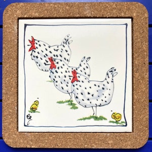 Puede incluir: Un azulejo de cerámica blanco con un diseño pintado a mano de tres gallinas moteadas de blanco y negro. Las gallinas están mirando en diferentes direcciones y tienen crestas rojas. Hay dos pollitos amarillos en la hierba debajo de las gallinas.