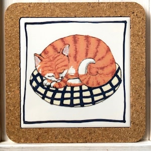 Könnte beinhalten: Ein Keramik-Untersetzer mit Korkrahmen zeigt eine handgemalte Illustration einer orangefarbenen Tabby-Katze, die auf einem blau-weiß karierten Kissen schläft.