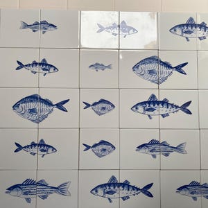 May include: A grid of square white ceramic tiles, each featuring a hand-painted blue illustration of a fish. The collection displays various fish species in different sizes and orientations, creating a repeating pattern across the wall surface.