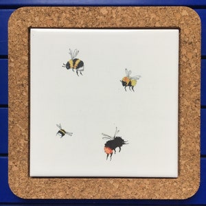 Puede incluir: Un azulejo de cerámica blanco con un marco de corcho presenta cuatro abejorros pintados a mano. Tres abejorros son amarillos y negros, y un abejorro es negro con acentos naranjas.