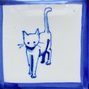 May include: Square ceramic tile featuring a blue cat illustration on a white background. The cat is depicted walking with its tail raised. The tile has a blue border.