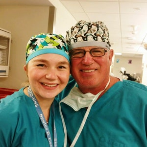 May include: Two people wearing teal scrubs and surgical caps. The person on the left is wearing a blue and green patterned cap with the text "SNHO-O/N Newton D. Fischer Soci..." on their lanyard. The person on the right is wearing a white and grey patterned cap with the text "DR. PALM" on the side.