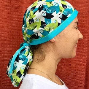 Puede incluir: Un gorro quir&uacute;rgico de color azul turquesa y blanco con un patr&oacute;n de b&uacute;hos con detalles en verde, negro y blanco. El gorro tiene un lazo de color azul turquesa.