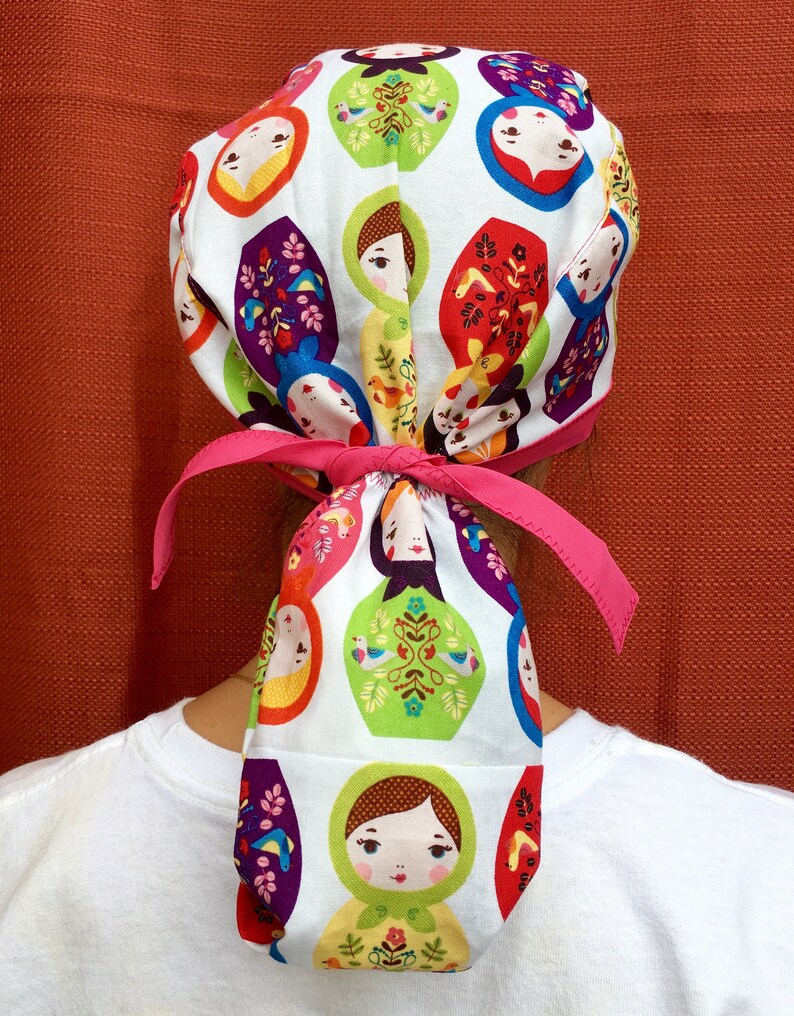 Puede incluir: Un gorro quir&uacute;rgico blanco con una cinta rosa, con un patr&oacute;n repetido de coloridas mu&ntilde;ecas rusas.
