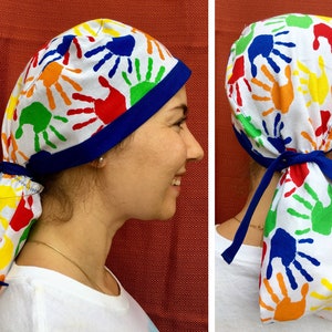 May include: A white surgical scrub hat with a blue trim and a colorful handprint pattern. The hat has a long tie that can be tied at the back of the neck.