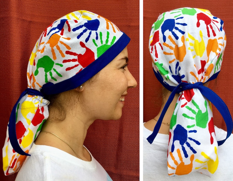 Puede incluir: Un gorro de quir&oacute;fano blanco con ribete azul y un estampado de huellas de manos de colores. El gorro tiene una larga corbata que se puede atar en la parte posterior del cuello.