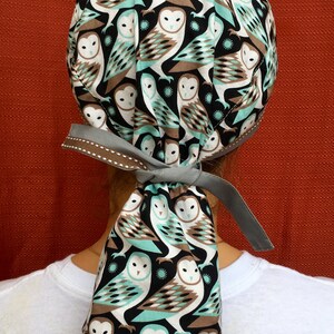 Puede incluir: Un gorro de quir&oacute;fano de tela con estampado gris y marr&oacute;n con una larga corbata. La tela presenta un patr&oacute;n repetido de b&uacute;hos blancos con detalles marrones y azul verdoso sobre un fondo negro.