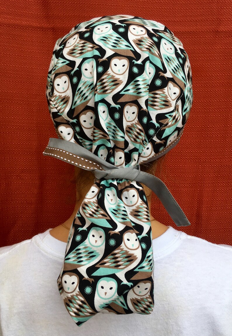 Puede incluir: Un gorro de quir&oacute;fano de tela con estampado gris y marr&oacute;n con una larga corbata. La tela presenta un patr&oacute;n repetido de b&uacute;hos blancos con detalles marrones y azul verdoso sobre un fondo negro.