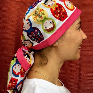 Puede incluir: Un gorro quir&uacute;rgico de color rosa y blanco con un lazo rosa. El patr&oacute;n presenta un dise&ntilde;o repetido de mu&ntilde;ecas rusas.