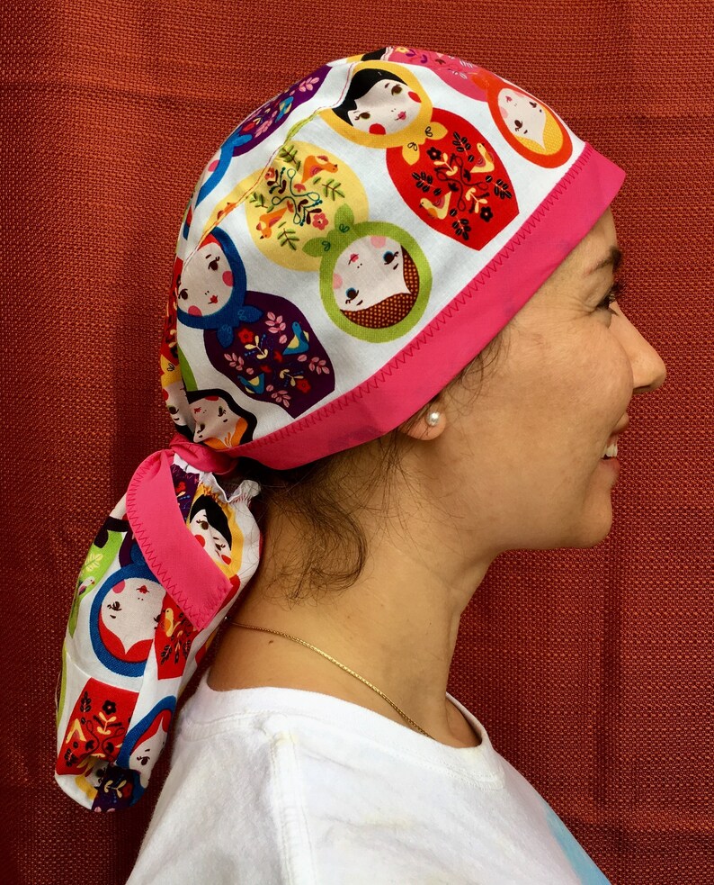 Puede incluir: Un gorro quir&uacute;rgico de color rosa y blanco con un lazo rosa. El patr&oacute;n presenta un dise&ntilde;o repetido de mu&ntilde;ecas rusas.