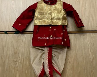 May include: A red and gold traditional Indian outfit for a child, consisting of a long-sleeved shirt, a waistcoat, and a dhoti. The shirt and waistcoat are decorated with gold embroidery and the dhoti is beige with red trim.