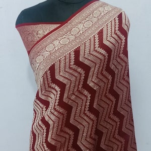 Op de afbeelding: Een rode chiffon sari met een gouden rand en een zigzagpatroon. De sari is gedrapeerd op een mannequin.