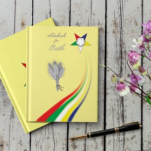 Peut inclure: Deux carnets jaunes avec une étoile à cinq branches et des épis de blé sur la couverture. Le carnet du dessus porte le texte "Notebook for Ruth". Un stylo noir et des fleurs roses sont sur une surface en bois.