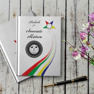 May include: White notebook with the text "Notebook for Associate Matron" and a five-pointed star with a sun symbol. The notebook is on a white wooden surface with pink flowers.