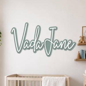 May include: A custom wall sign featuring the name Vada Jane in a layered script font. The sign consists of a sage green backing layer with a white top layer, creating a dimensional effect. The letters are connected in a cursive style.