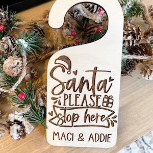 Könnte beinhalten: Ein hölzerner Türhänger mit dem Text "Santa Please Stop Here" und den Namen "Maci & Addie" darunter. Der Hänger ist mit einem Weihnachtsmann-Hut und einer Geschenkbox verziert.