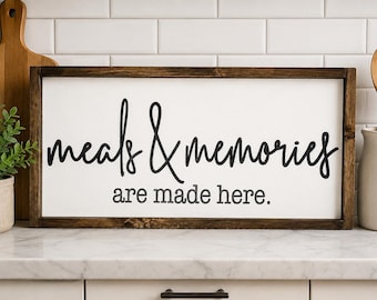 May include: A rectangular wooden sign with a white background and a dark brown frame. The sign reads "meals & memories are made here" in black cursive lettering. Kitchen decor.
