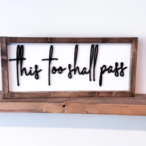 May include: A rectangular wooden sign with the phrase "This too shall pass" in black cursive lettering. The sign has a white background and a dark brown wooden frame. It is displayed on a wooden shelf.