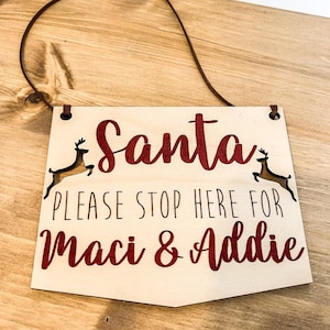 May include: A wooden sign with a red string for hanging. The sign says "Santa Please Stop Here For Maci & Addie" in red and black lettering. There are two reindeer cutouts on either side of the sign.