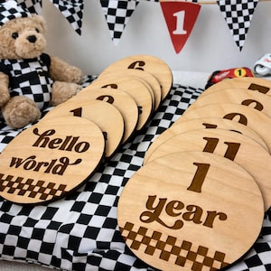 以下が含まれることがあります： 「hello world」と「1 year」を含む、数字とテキストが刻まれた木製のマイルストーンディスク。 ディスクは、黒と白のチェック柄の表面に配置され、背景にはテディベアとチェック柄の旗のバナーがあります。 赤いペナントには数字の1が表示されています。
