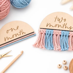 Puede incluir: Marcadores de hitos de madera con flecos de macramé rosas y azules, que indican "un mes" y "dos meses". La imagen incluye ovillos de lana, tijeras, un ganchillo y cuentas de madera, sugiriendo un tema artesanal.