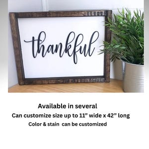 May include: A wooden sign with a dark brown frame and a white background. The word "thankful" is written in black cursive lettering. The sign is available in several sizes up to 11 inches wide by 42 inches long. The color and stain can be customized.