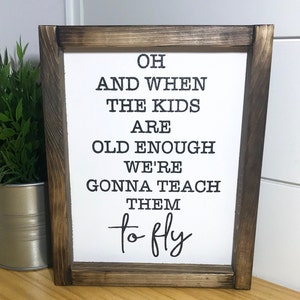 May include: A framed sign with a white background and black text that reads "Oh and when the kids are old enough we're gonna teach them to fly". The frame is made of dark brown wood.