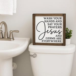 May include: A framed wooden sign with the text "WASH YOUR HANDS AND SAY YOUR PRAYERS Jesus AND GERMS ARE EVERYWHERE" in black script. The sign is displayed on a white toilet, next to a small potted plant. A white sink and faucet are visible.