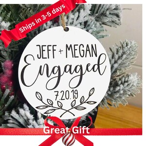 May include: White round bauble with black calligraphy that reads "Jeff + Megan Engaged 7.20.19" with a floral design. The bauble is hanging on a snowy evergreen tree branch.