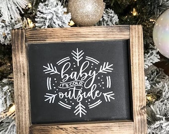 Può includere: Un cartello in legno rustico con cornice in legno e lavagna nera su cui è scritto il testo "baby it's cold outside" in lettere bianche in corsivo. Il cartello è decorato con un motivo a fiocco di neve.