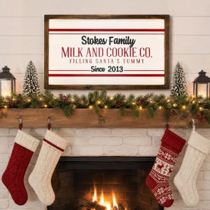 Puede incluir: Un letrero rectangular de madera con fondo blanco y marco de madera oscura. El texto dice Stokes Family MILK AND COOKIE CO. FILLING SANTA'S TUMMY Since 2013 en letras negras y rojas, acentuado por líneas horizontales de color rojo.