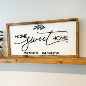 May include: A white wooden sign with a brown frame, featuring the text "Home Sweet Home" in black cursive script with a small leaf design. The sign also includes the coordinates "33.9161°N -84.4122°W" in black text.