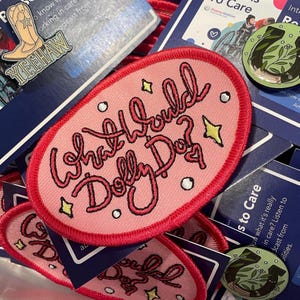 May include: An oval-shaped embroidered patch with a pink background and a red border. The text "What Would Dolly Do?" is stitched in dark pink cursive lettering. The design includes small yellow four-pointed stars, white circles, and a small heart.