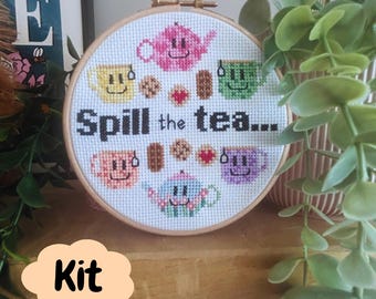 May include: A circular wooden embroidery hoop containing a cross-stitch pattern on white fabric. The design features two teapots, four smiling teacups, and various cookies, with the black text "Spill the tea..." centered in the middle.